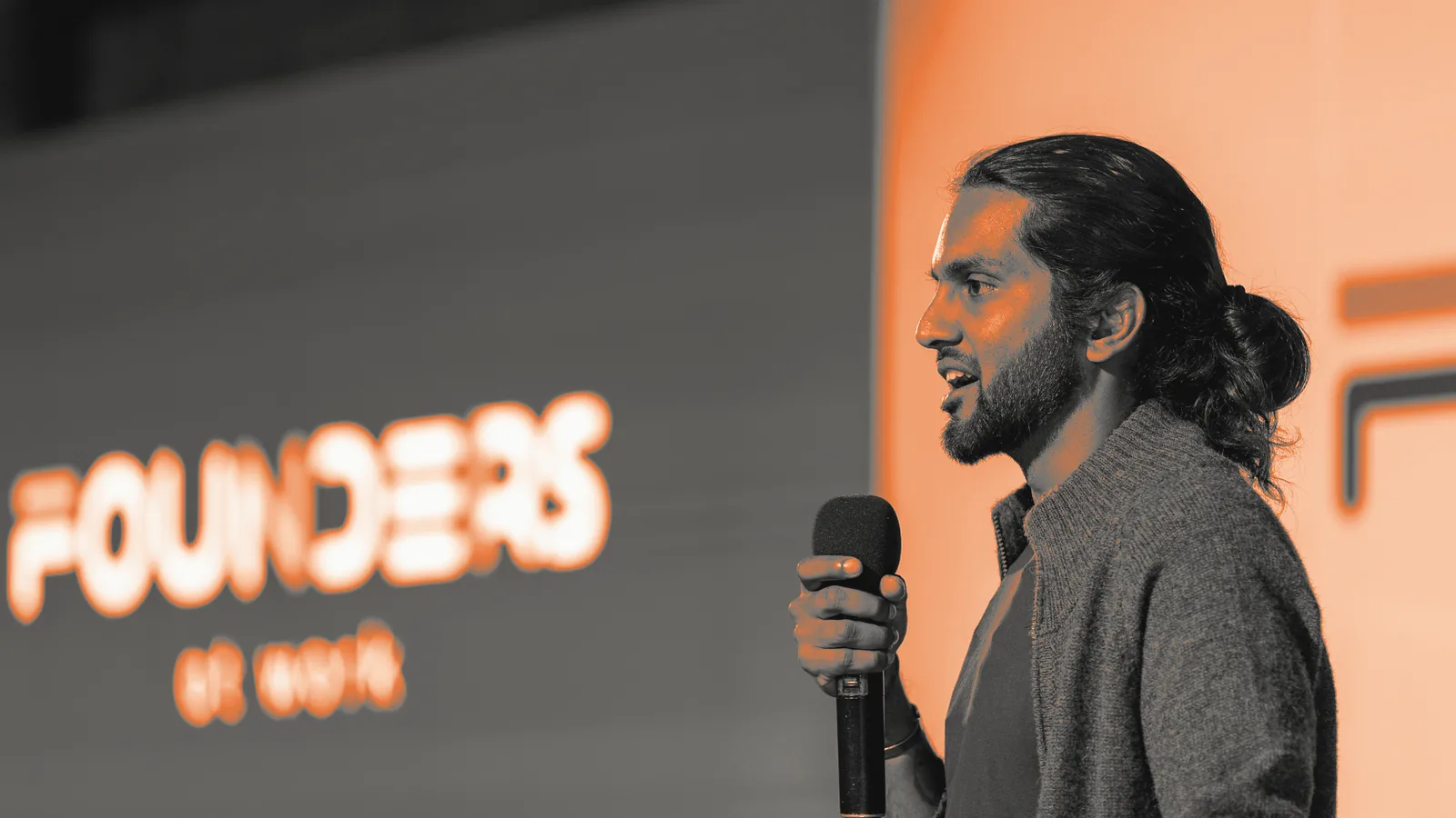 Keshav Mohabir speaking with a microphone on the Founders at Work Summit stage, the event logo on the screen behind him